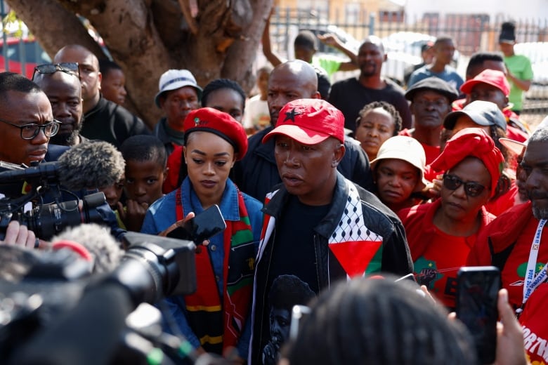 A crowd of people is shown outdoors, with people seeming to surround a man in a baseball cap and a woman in a beret.