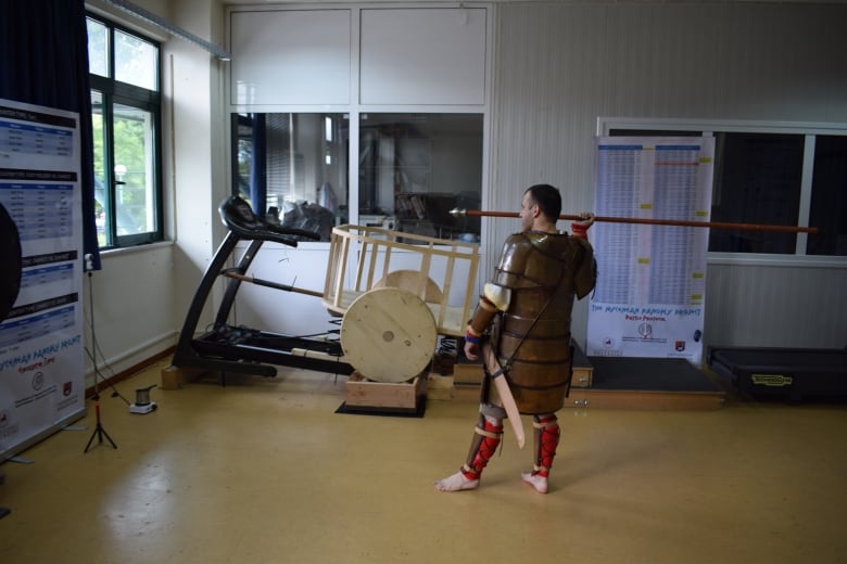 A barefoot man in a full suit of plated bronze armour stands in a modern room next to a treadmill holding a javelin over his shoulder.