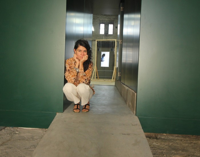 A young woman with dark hair crouches in a gallery space, facing the camera