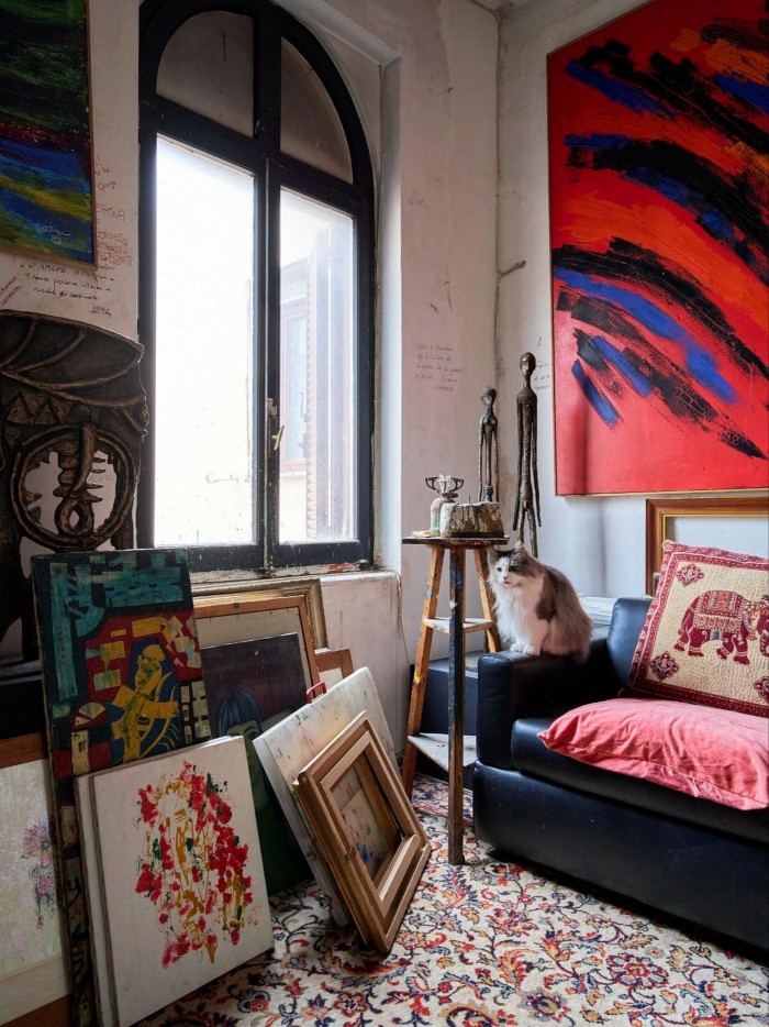 A photograph of the interior of an artist’s studio with a large window, framed paintings propped up on the floor — and a cat perched on a sofa