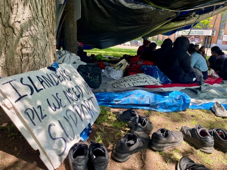 A sign that reads "Islander plz we need your support" in the foreground of a tent encampment.