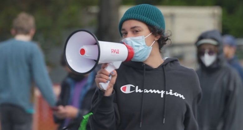 A person in a green toque, a blue medical mask and a black hoodie holds a loudspeaker up to their face. 