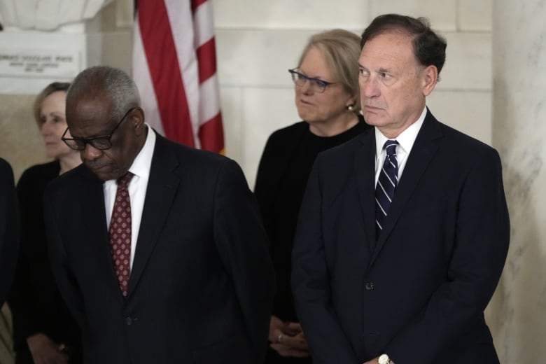 Two men in suits, one dark complected, and a woman wearing glasses are shown standing in an indoor photo. All are wearing dark clothing.