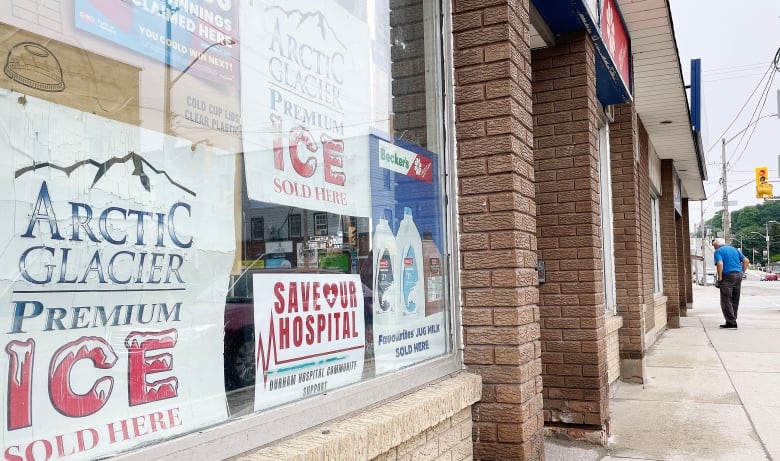 A sign that says "save our hospital" is in the window of a store with advertisements for ice and milk beside it.