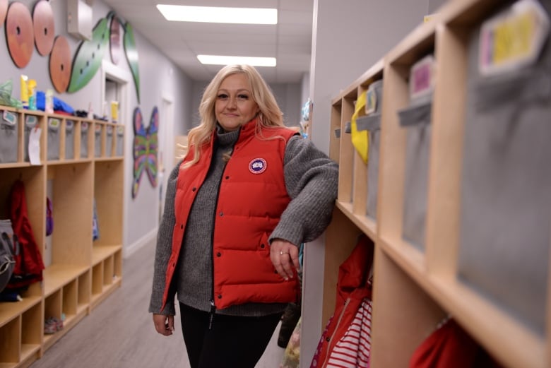 A woman with long, blond hair wearing a red puffer vest and a grey sweater stands in the hallway of a daycare centre among toy bins and colourful wall art. 