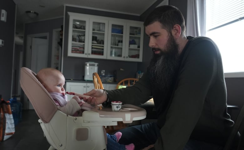 A baby girl is sitting in a high chair, while her father fits in front of her and feeds her mashed fruit.