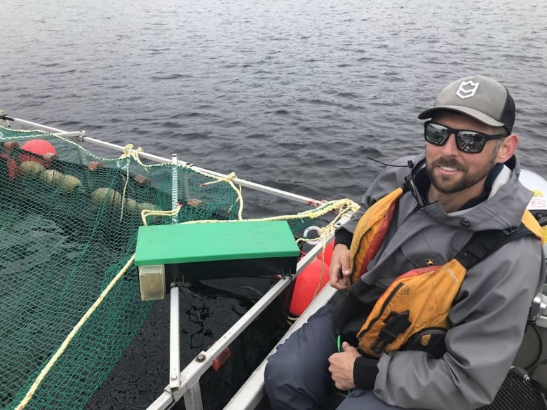 A man wearing a life jacket, a baseball cap, and a pair of sunglasses, sits on a boat. A net and rope can be seen to his left. 