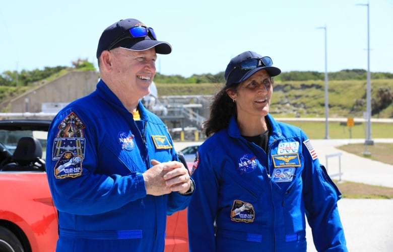 Two astronauts wearing blue jumpsuits and baseball caps stand smiling.