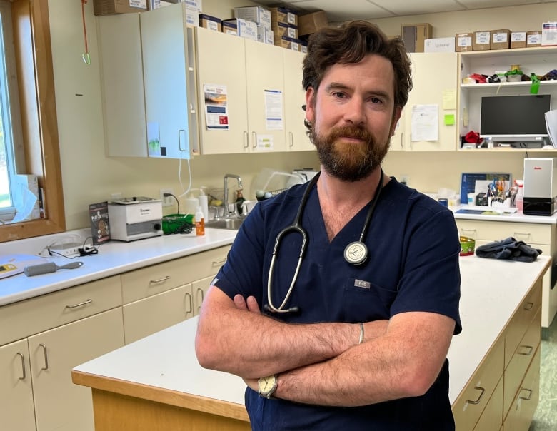 A man with reddish brown curly hair and a beard wearing a dark blue shirt and a stethoscope stands with arms crossed in front of a wall of cabinets.  