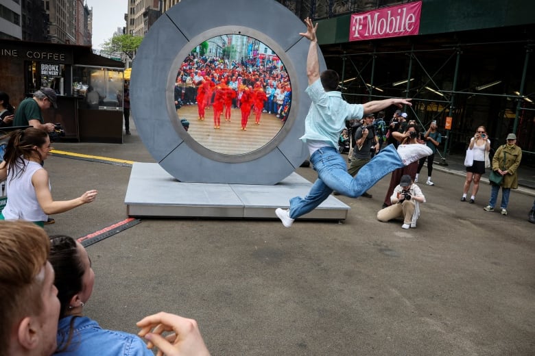 A man leaps in a dramatic dance movement in front of a large circular screen in the street showing a live feed of a group of dancers all dressed in bright orange. 