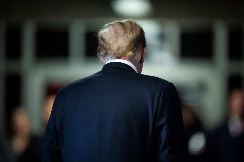 A man with thin grey hair wearing a blue suit walks away from a crowd of reporters.