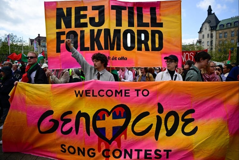 Protesters gather while holding up a banner that reads, 'Welcome to genocide song contest.'