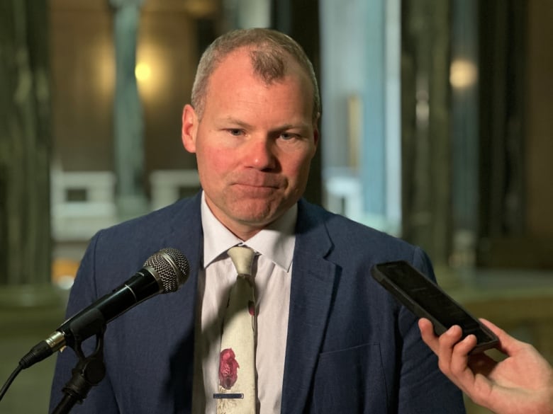 A white man with thinning grey hair is wearing a suit and tie. He has a neutral look on his face.