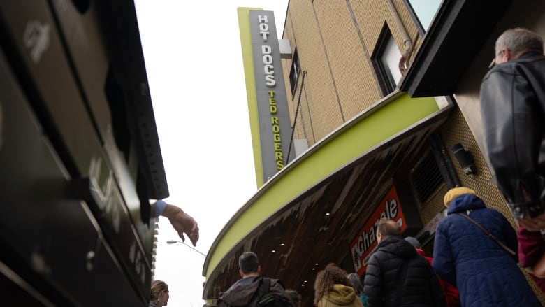 A lineup of people stand in front of a theatre. A sign above them reads "Hot Docs Ted Rogers"