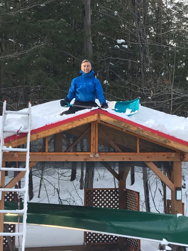 A man sits on a snowy roof with a shovel.