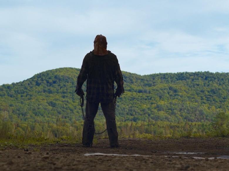 A man wearing a mask and holding a chain looks out at a forested hill.