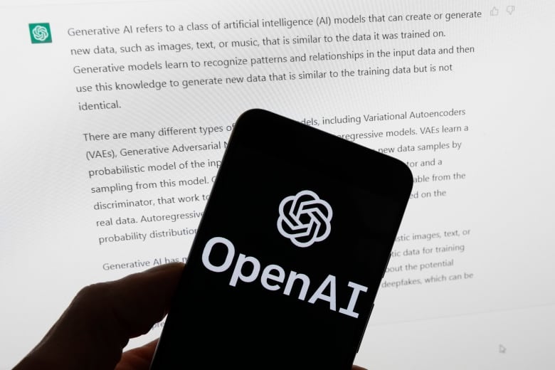 A person holds a phone displaying the word, 'OpenAI,' in front of a computer screen displaying text.