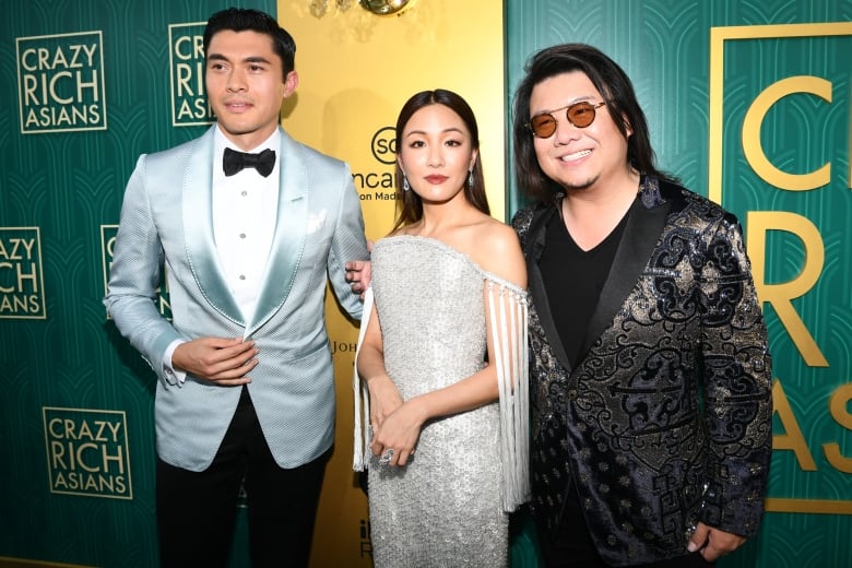 A woman in a white gown stand between two men in silk suits in front of a wall with the words "Crazy Rich Asians."