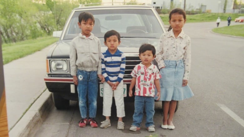 Four kids stand in front of a blue car.