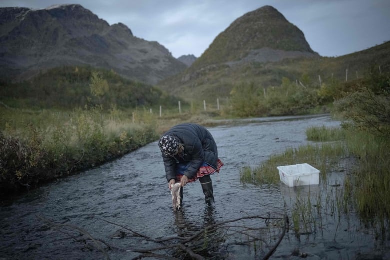 A woman leans over a stream with hills in the background