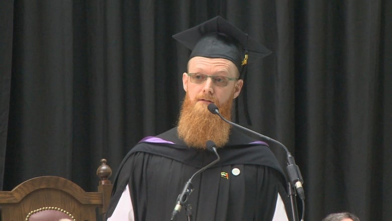 A man wearing a graduation cap and gown speaking in front of a microphone.