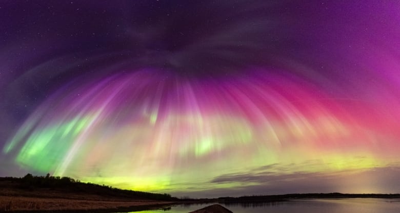 A beautiful display of northern lights taken at night looks like it's forming an umbrella in the way the charged particles are streaming down over a body of water in a very flat landscape, The colours are spectacular, starting with violet at the top of the umbrella, then turning into hot pink, magenta, citrus yellow, vibrant green and a bit of orange at the bottom. 