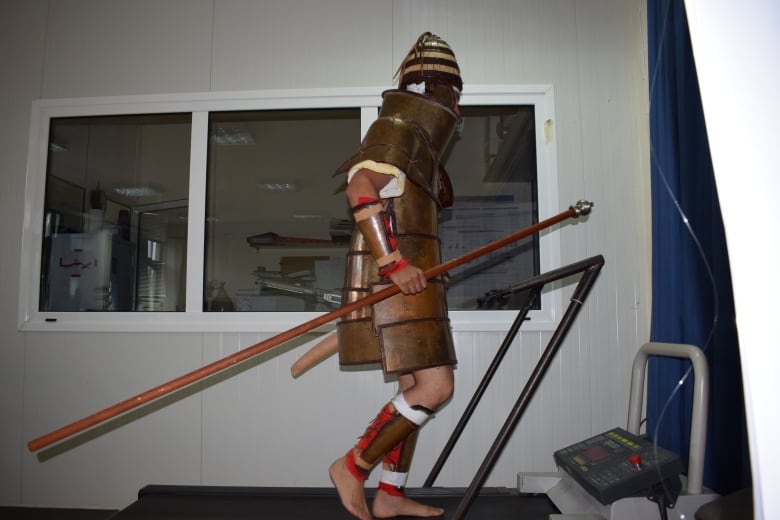 A man in a full suit of bronze armour, pictured in profile against a black backdrop as he lunges a spear into a wooden shield. 