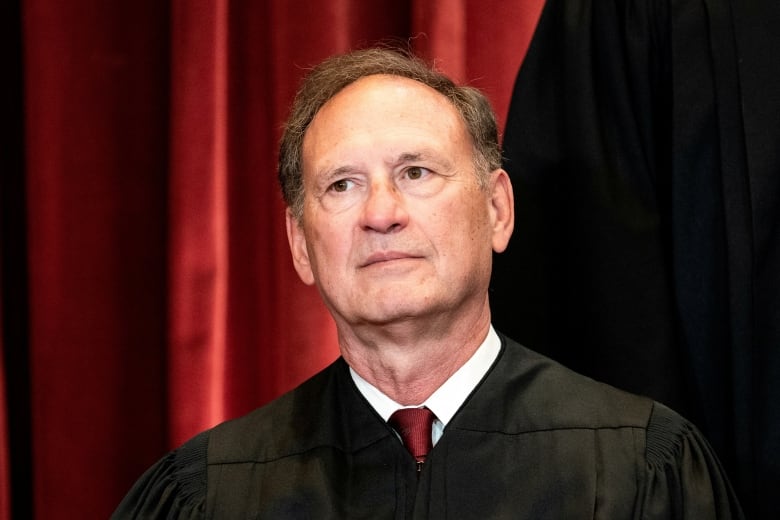 An older cleanshaven man in a tie and judicial robes is shown in closeup.