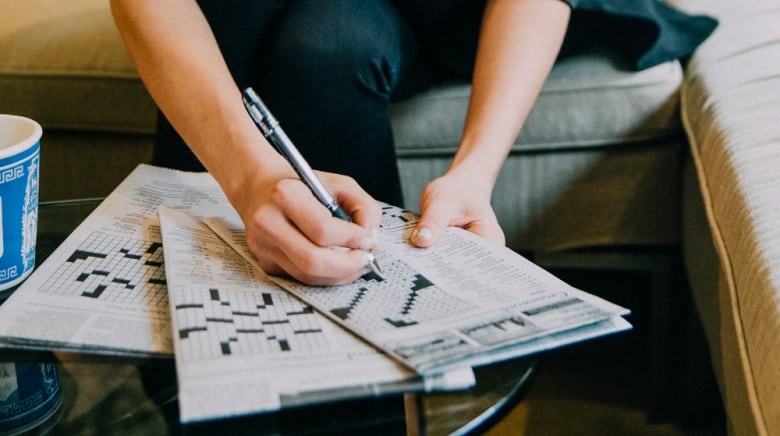 A woman with shoulder-length hair in a button-up shirt is sitting on couch looking down at a newspaper on a coffee table in front of her. The newspaper is open to the crossword puzzle and she is writing on the page with a pen. 