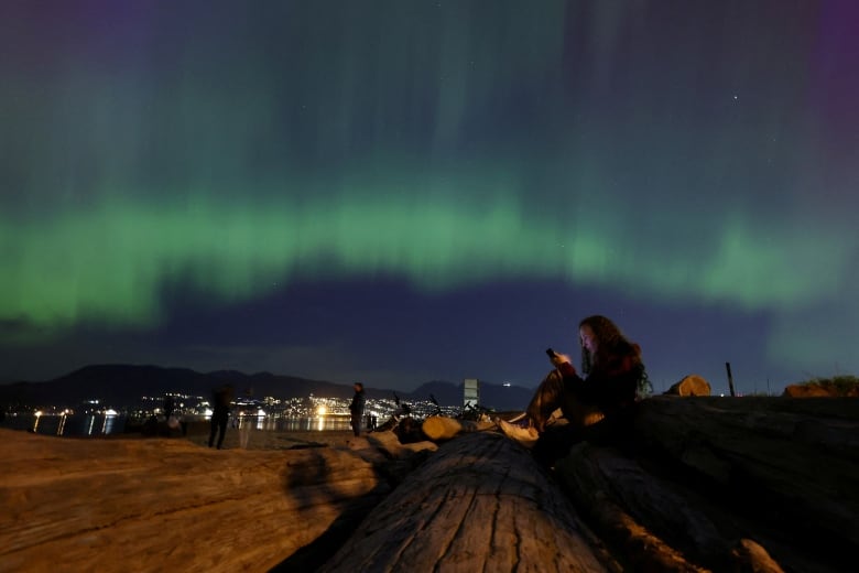 A person checks their phone while seated on a log, as the northern lights illuminate the sky above them.