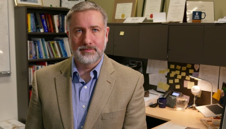 A man in a beige suit jacket with blue shirt is standing in his office with books and cabinets behind him.