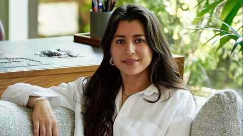 Portrait of a smiling woman with dark, shoulder-length hair, dressed in a casual white shirt, relaxing in a home environment filled with natural light and lush greenery