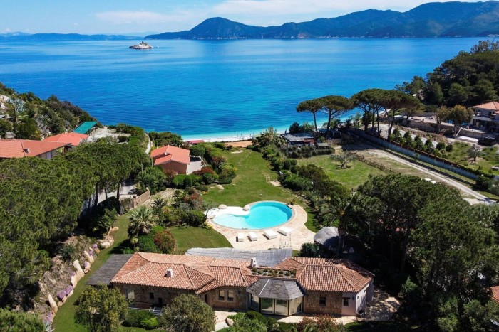 aerial view of a villa with a kidney-shaped pool, near the waterfront of a bay surrounded by lush greenery