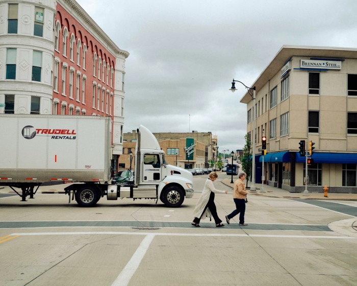 Two people cross a road in a downtown area. Behind them a large truck passes the junction