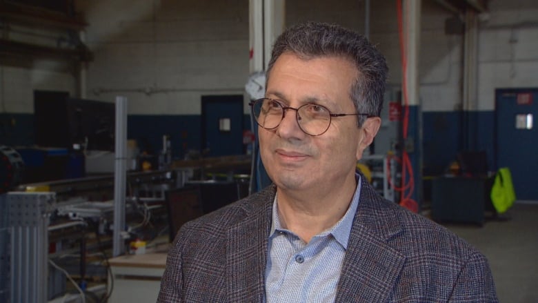 A man in glasses and knit blazer smiles slightly off-camera toward the left as he stands indoors in a factory setting.