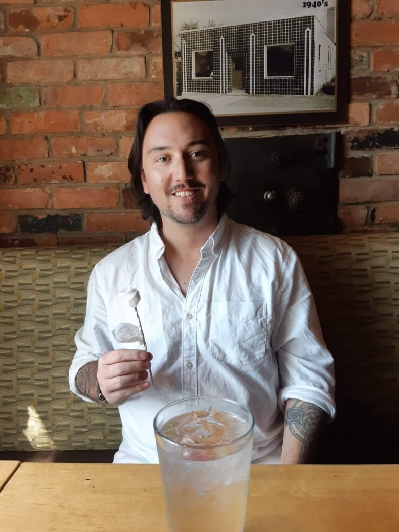 A smiling man holding a white paper rose sits at a restaurant table. There is a glass filled with ice and water in front of him. 