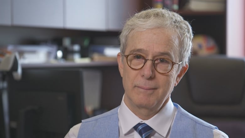 Man with white hair, round glasses and a blue tie and vest. 