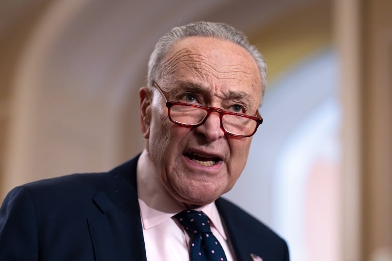 A white man wearing a suit and red-tinged eyeglasses speaks in a large building.