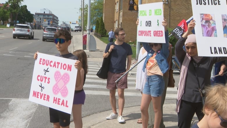 A crowd of half a dozen people hold signs on a city sidewalk. In the foreground, a boy holds a sign that reads "Education cuts never heal"