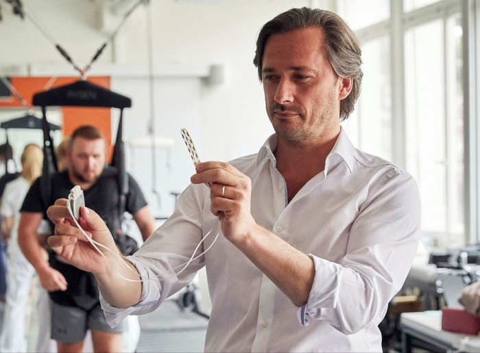 Gregoire Courtine wearing a white shirt holds up the EPFL implant. A man suspended from a harness is in the background