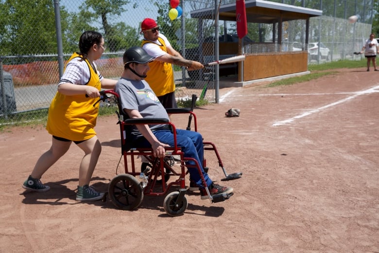 A man in a wheelchair is at home plate of a baseball field, and is being pushed by a woman wearing a bright yellow jersey to run the base.