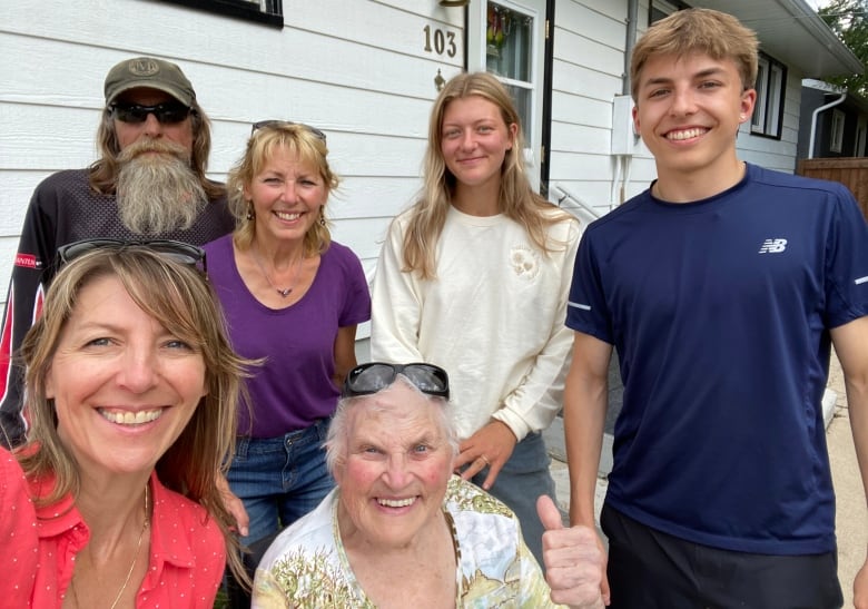 Woman with grey hair sits in the middle. From left blond woman with pink shirt, man with long beard and baseball hat, blond woman with purple shirt, blond woman with white shirt and blond young man with blue shirt surround her. 