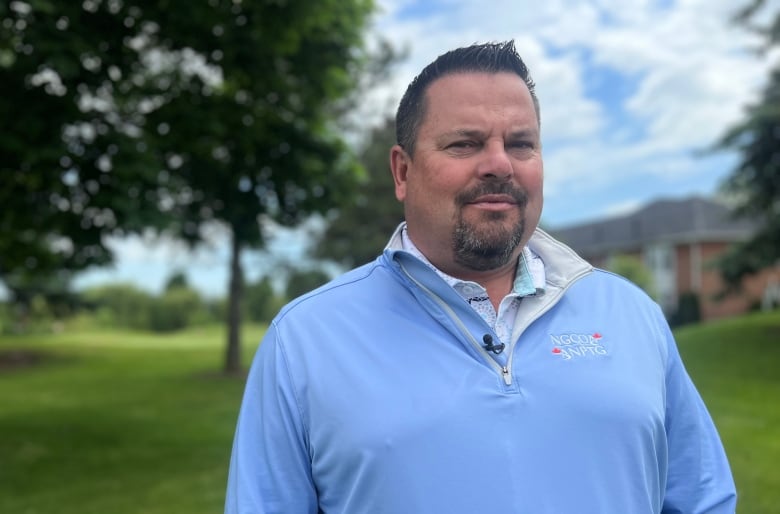 A man is standing in front of a golf course wearing a light blue shirt. 