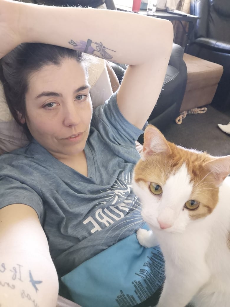 A woman with dark hair lies on a couch, a heating pad and tabby cat on her lap.