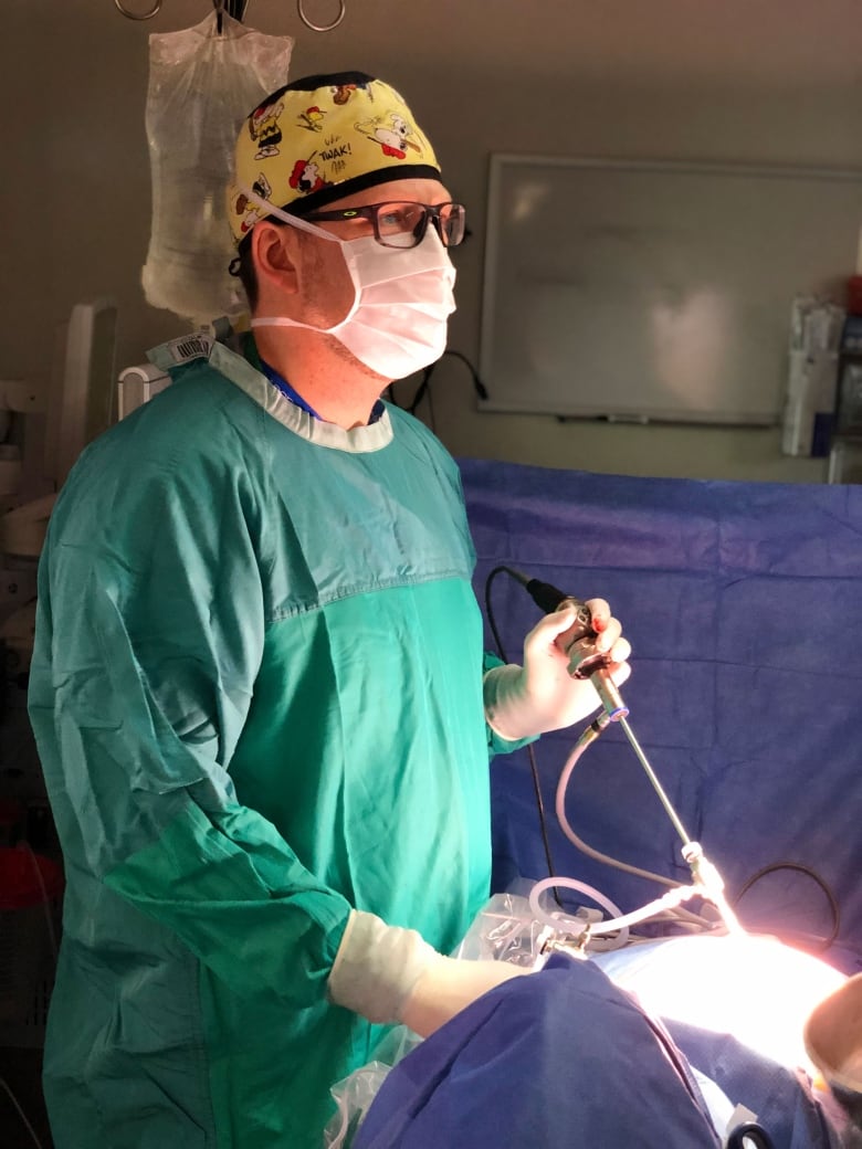 A surgeon wearing a green gown, yellow cap with Peanuts cartoons on it, a medical mask and gloves holds a long instrument in an operating room.