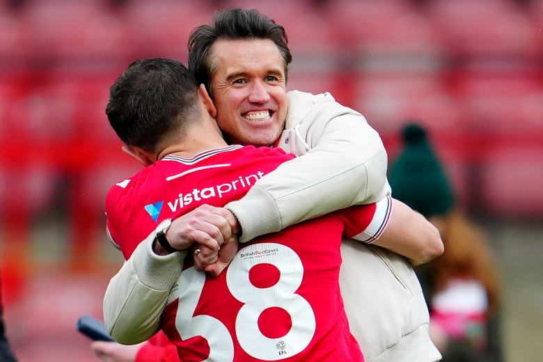 A man wearing a white sports jacket hugs a football player wearing a red jersey and number 38.