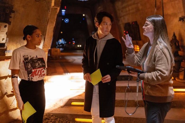 Three people stand in a room. The two standing on the left hold yellow paper in their hands. The person standing on the right holds an ipad and is speaking. 