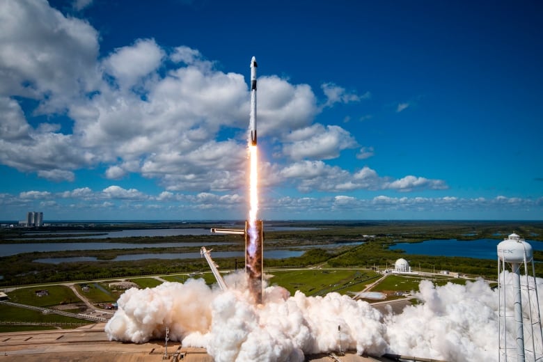 A white rocket leaves a trail of smoke on a launch pad as it soars into a blue sky dotted by white clouds.