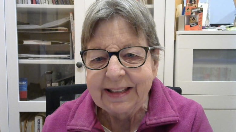 A woman with grey hair and glasses is interviewed in front of a white cabinet housing papers and books.