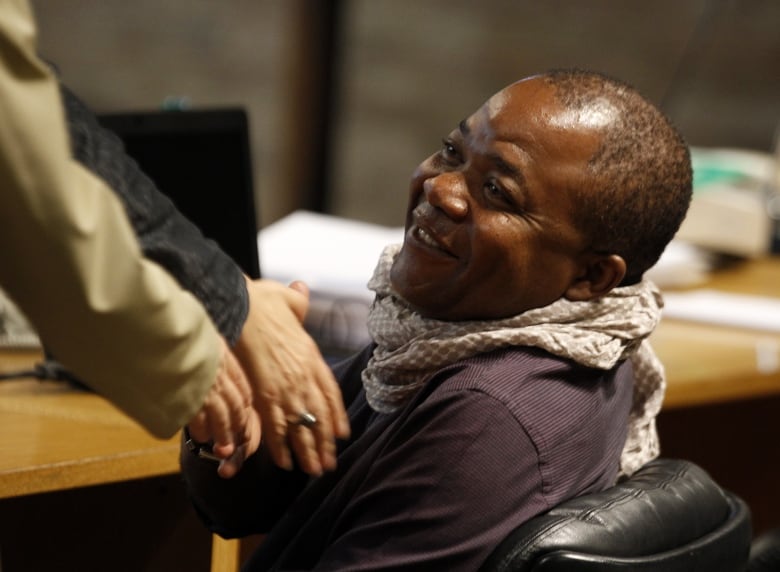 A smiling darkskinned, clean shaven man is shown sitting at a table indoors.